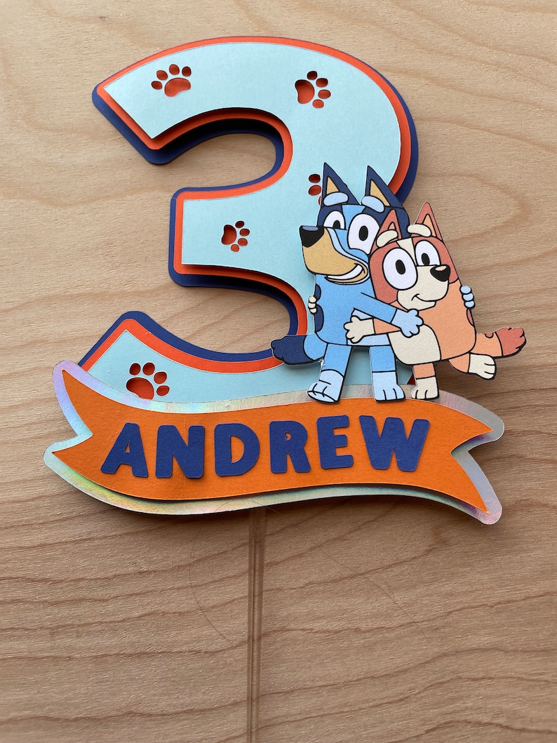 Puede incluir: Un adorno para pastel 3D azul y naranja con el n&uacute;mero 3, huellas de patas y los personajes del programa Bluey. El adorno tambi&eacute;n incluye el nombre Andrew en una pancarta.
