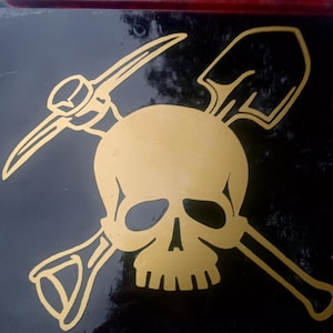 May include: A gold skull with a pickaxe and shovel crossed behind it. The skull is the focal point of the image.