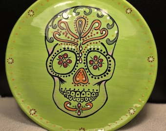 May include: A green ceramic plate with a painted sugar skull design. The skull is decorated with black, orange, red, and white details. The plate has a white rim with orange dots.