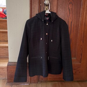 May include: A black hooded jacket featuring a vertical ribbed texture, a front button closure with silver-toned hardware, and two horizontal zippered pockets at the waist. The jacket includes a drawstring hood with silver aglets and a visible inner lining.