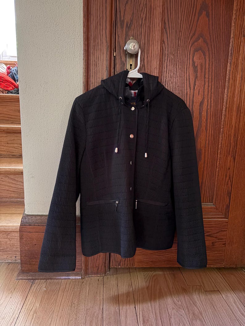 May include: A black hooded jacket featuring a vertical ribbed texture, a front button closure with silver-toned hardware, and two horizontal zippered pockets at the waist. The jacket includes a drawstring hood with silver aglets and a visible inner lining.