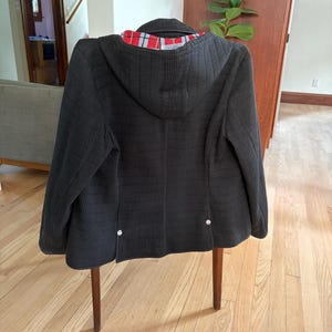 May include: A black hooded jacket with a textured, ribbed exterior surface. The interior lining features a red, white, and black plaid pattern. The back of the jacket includes two small silver-toned metal buttons near the bottom hemline.