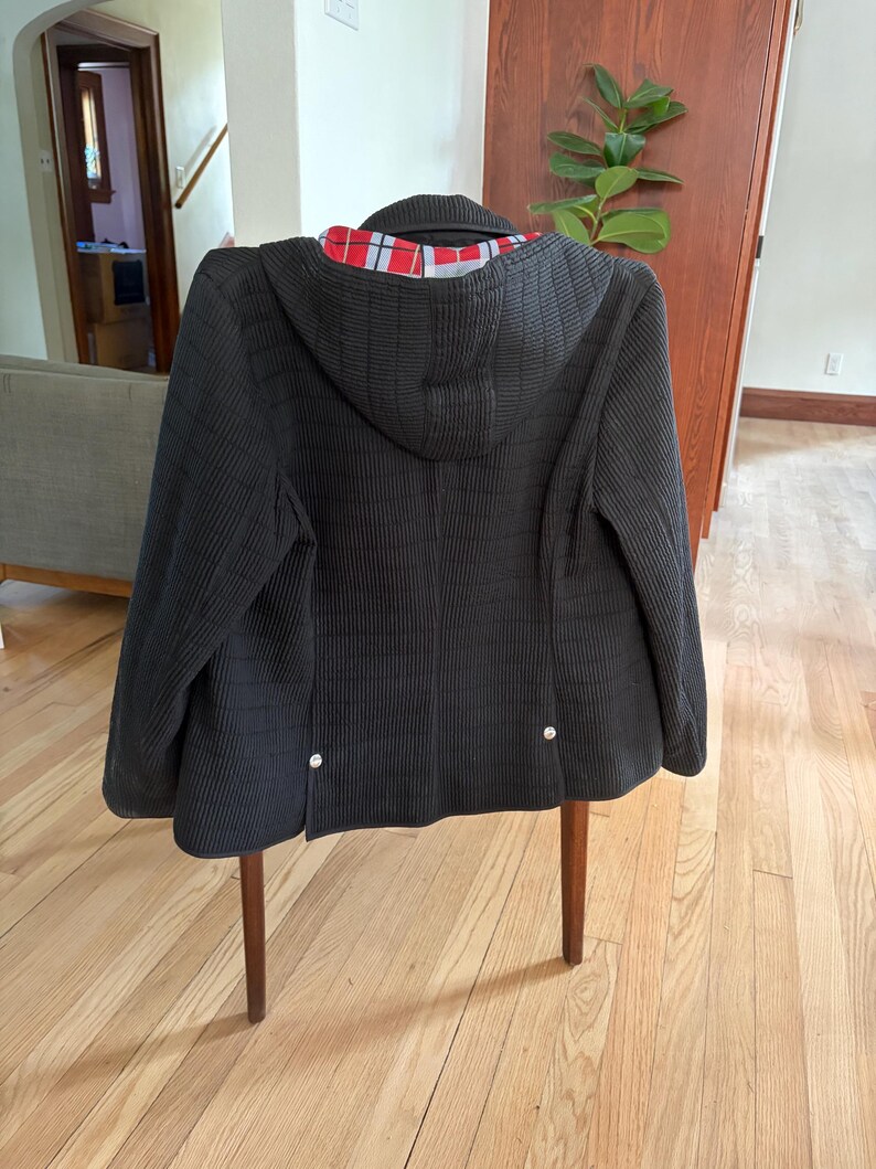 May include: A black hooded jacket with a textured, ribbed exterior surface. The interior lining features a red, white, and black plaid pattern. The back of the jacket includes two small silver-toned metal buttons near the bottom hemline.