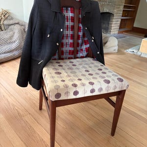 May include: A wooden chair with a beige seat cushion featuring a repeating pattern of dark purple circles. A black hooded jacket with a red and white plaid interior lining is draped over the backrest of the chair, showing silver-toned hardware.
