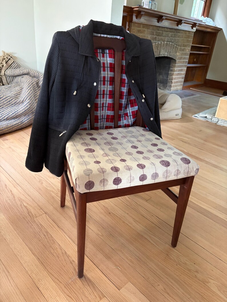 May include: A wooden chair with a beige seat cushion featuring a repeating pattern of dark purple circles. A black hooded jacket with a red and white plaid interior lining is draped over the backrest of the chair, showing silver-toned hardware.