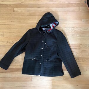 May include: A black quilted jacket featuring a hood with a red, white, and blue patterned lining. The jacket has long sleeves, a front button closure with silver-toned buttons, and two zippered pockets on the lower front section.