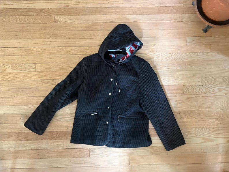 May include: A black quilted jacket featuring a hood with a red, white, and blue patterned lining. The jacket has long sleeves, a front button closure with silver-toned buttons, and two zippered pockets on the lower front section.
