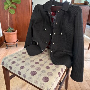 May include: A black quilted jacket with a hood, featuring a front zipper closure, silver-toned metal snaps, and two zippered pockets. The interior lining displays a red, black, and white plaid pattern, and the jacket has a ribbed texture.