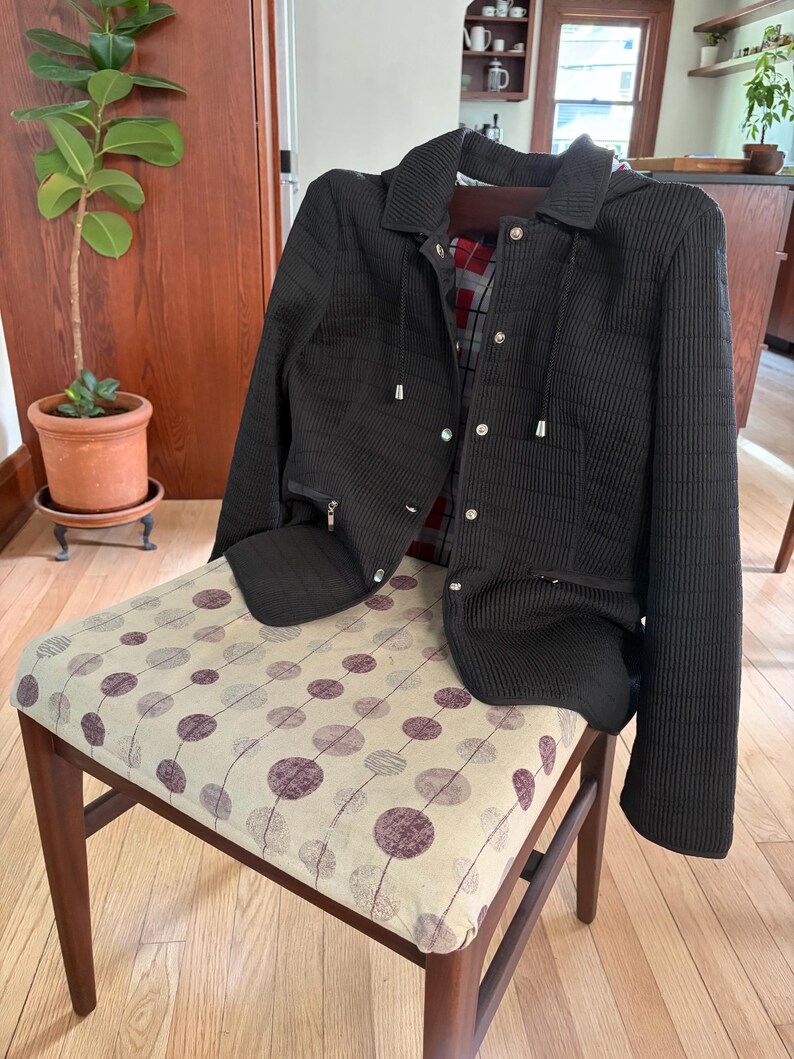 May include: A black quilted jacket with a hood, featuring a front zipper closure, silver-toned metal snaps, and two zippered pockets. The interior lining displays a red, black, and white plaid pattern, and the jacket has a ribbed texture.