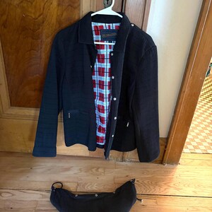 May include: A black corduroy jacket with a fold-down collar, long sleeves, and a front button closure. The interior features a red, white, and blue plaid lining with a Pendleton label. A matching black corduroy hood is positioned below the jacket.