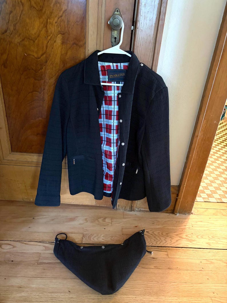 May include: A black corduroy jacket with a fold-down collar, long sleeves, and a front button closure. The interior features a red, white, and blue plaid lining with a Pendleton label. A matching black corduroy hood is positioned below the jacket.