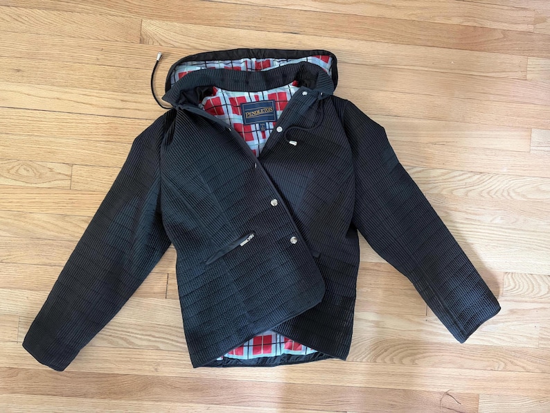 May include: A black quilted jacket with a hood and a red, white, and blue plaid lining. The jacket features a front zipper closure with silver-tone metal snaps, long sleeves, and a Pendleton brand label sewn into the interior neck area.