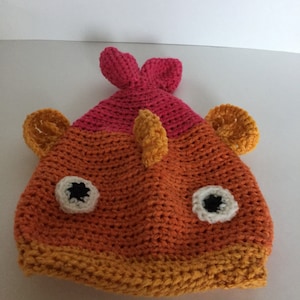 May include: A crocheted hat shaped like a fish with orange, yellow, and pink yarn. The hat has two large ears, a fin on top, and two white eyes with black pupils.