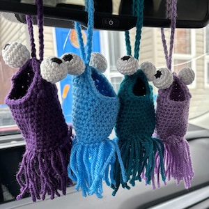 May include: Five crocheted hanging ornaments in the shape of a monster with large eyes and fringe. The ornaments are purple, blue, teal, and lavender.