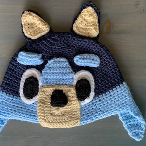 May include: A blue and beige crocheted hat shaped like a dog's head with black eyes and a black nose.