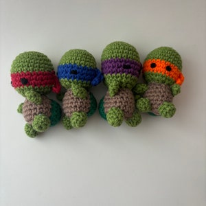 May include: Four crocheted Teenage Mutant Ninja Turtles, each with a different coloured mask: red, blue, purple, and orange. The turtles are green and brown and have black eyes.