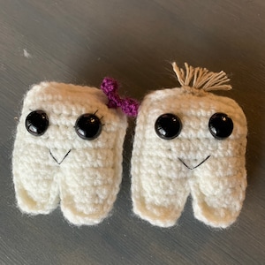 May include: Two crocheted tooth-shaped plushies with black eyes and a small purple bow on one. The plushies are white and have a crocheted texture.