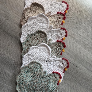 May include: A collection of handmade crochet chicken-shaped coasters in various colours. The coasters are white, brown, and green, with red and yellow accents. The coasters are arranged on a grey wooden surface.