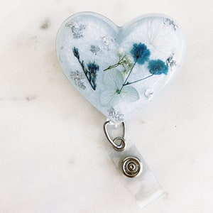 May include: A white heart-shaped resin keychain with a blue and white pressed flower design and silver leaf accents. The keychain has a clear plastic retractable badge holder.