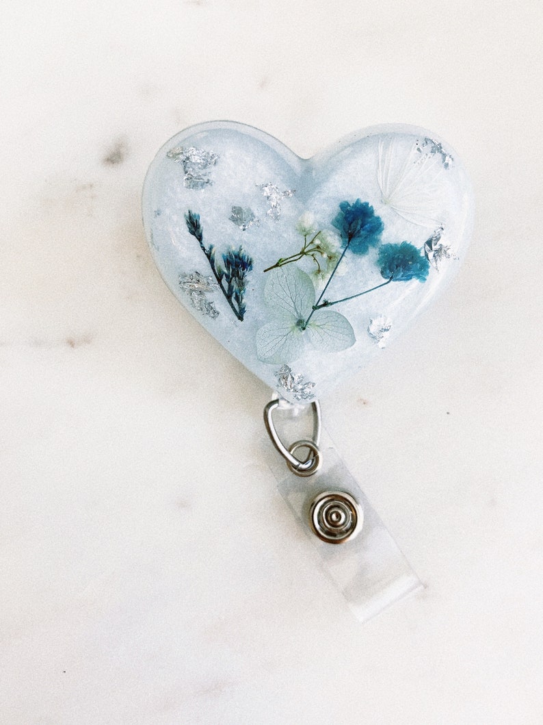 May include: A white heart-shaped resin keychain with a blue and white pressed flower design and silver leaf accents. The keychain has a clear plastic retractable badge holder.