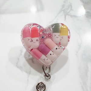 May include: A pink heart-shaped resin keychain with a clear background and glitter. The keychain features five colorful pill charms with cute faces. The keychain is attached to a clear plastic badge holder with a silver clip.