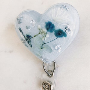 May include: A heart-shaped resin badge holder with a clear background and blue and white pressed flowers. The holder is attached to a clear retractable badge reel with a silver clip and button.