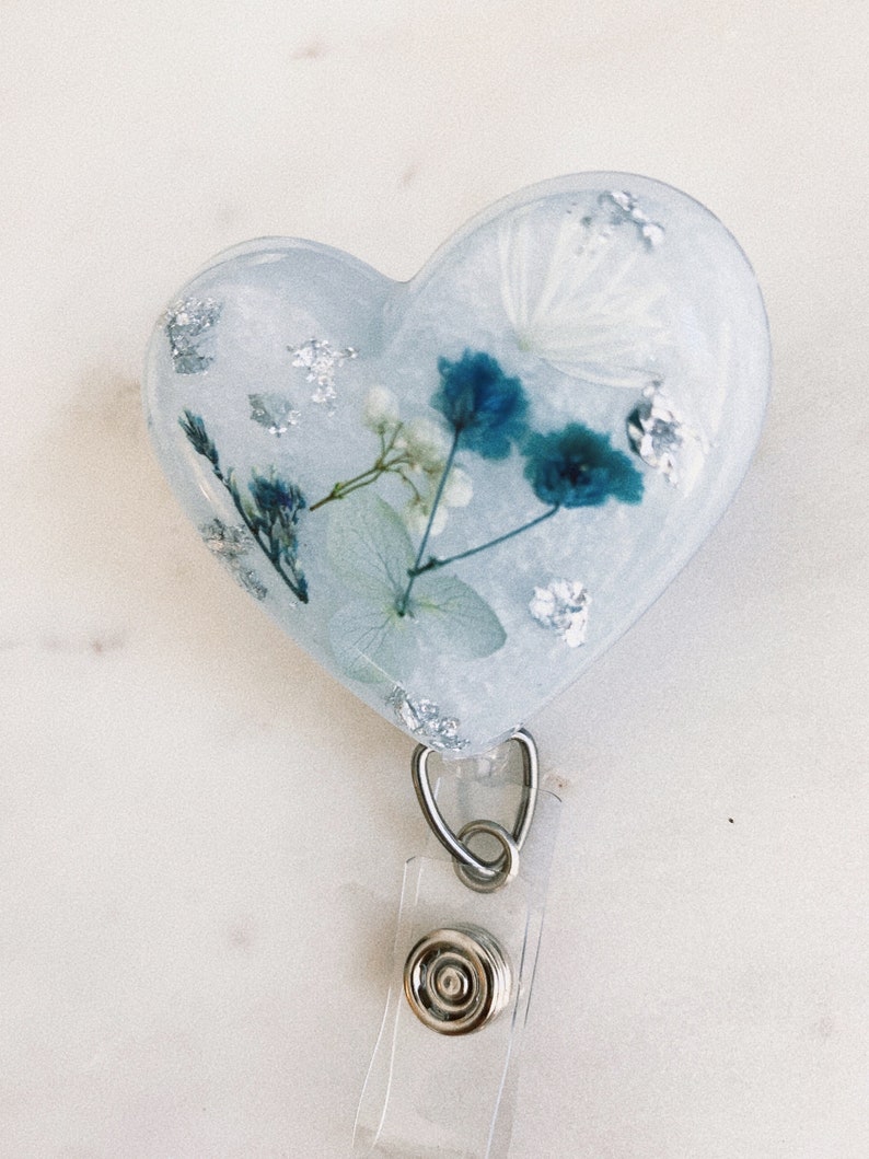May include: A heart-shaped resin badge holder with a clear background and blue and white pressed flowers. The holder is attached to a clear retractable badge reel with a silver clip and button.