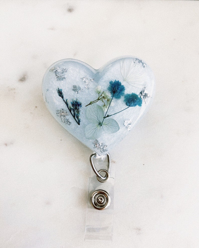 May include: A heart-shaped resin badge holder with a white background and blue and white pressed flowers. The holder has a clear plastic retractable badge clip.