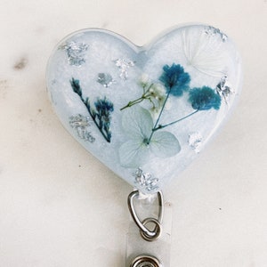 May include: A heart-shaped resin badge holder with a blue and white pressed flower design. The holder has a silver glitter finish and a clear plastic retractable badge clip.