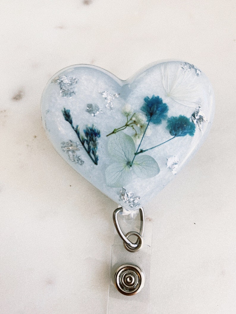 May include: A heart-shaped resin badge holder with a blue and white pressed flower design. The holder has a silver glitter finish and a clear plastic retractable badge clip.