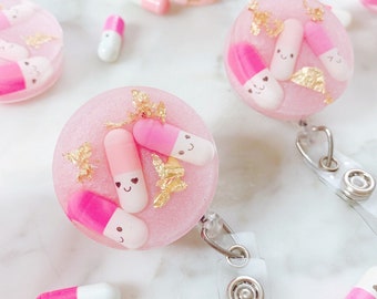 May include: Pink and white pill shaped resin badge holder with gold glitter and gold leaf accents. The badge holder has a clear plastic strap with a silver clip.