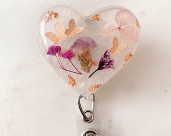 May include: Heart-shaped badge reel with a clear resin design. The resin encases dried flowers in shades of pink and purple, along with gold butterfly accents and gold leaf flakes. The reel has a silver clip and a clear plastic strap.