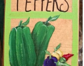 Op de afbeelding: Een houten bord met het woord "PEPPERS" in zwart op een groene achtergrond. Het bord heeft een schilderij van twee groene paprika's, een paarse paprika, een gele paprika en een rode paprika. De handtekening van de kunstenaar "Shari" is in rood onderaan het bord geschilderd.