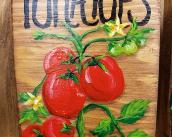 Op de afbeelding: Een houten bord met het woord "TOMATOES" in zwarte letters. Het bord heeft een geschilderd beeld van een tomatenplant met rode tomaten, groene bladeren en gele bloemen.