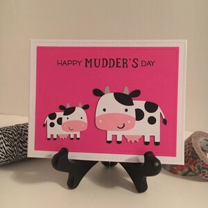 May include: A pink card with the text "Happy Mudder's Day" and two black and white cows. The card is on a black stand.