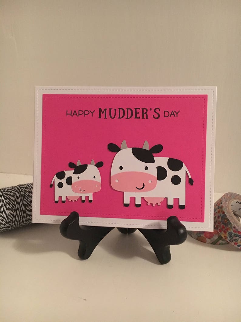 May include: A pink card with the text "Happy Mudder's Day" and two black and white cows. The card is on a black stand.