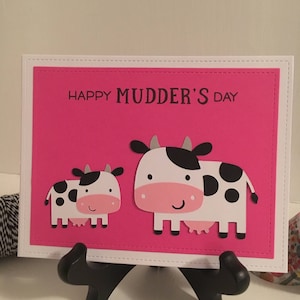May include: A pink card with a white border and the text "Happy Mudder's Day". Two white cows with black spots are on the card. The larger cow is on the right and the smaller cow is on the left.