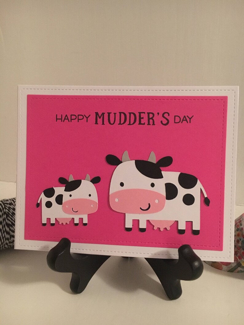 May include: A pink card with a white border and the text "Happy Mudder's Day". Two white cows with black spots are on the card. The larger cow is on the right and the smaller cow is on the left.