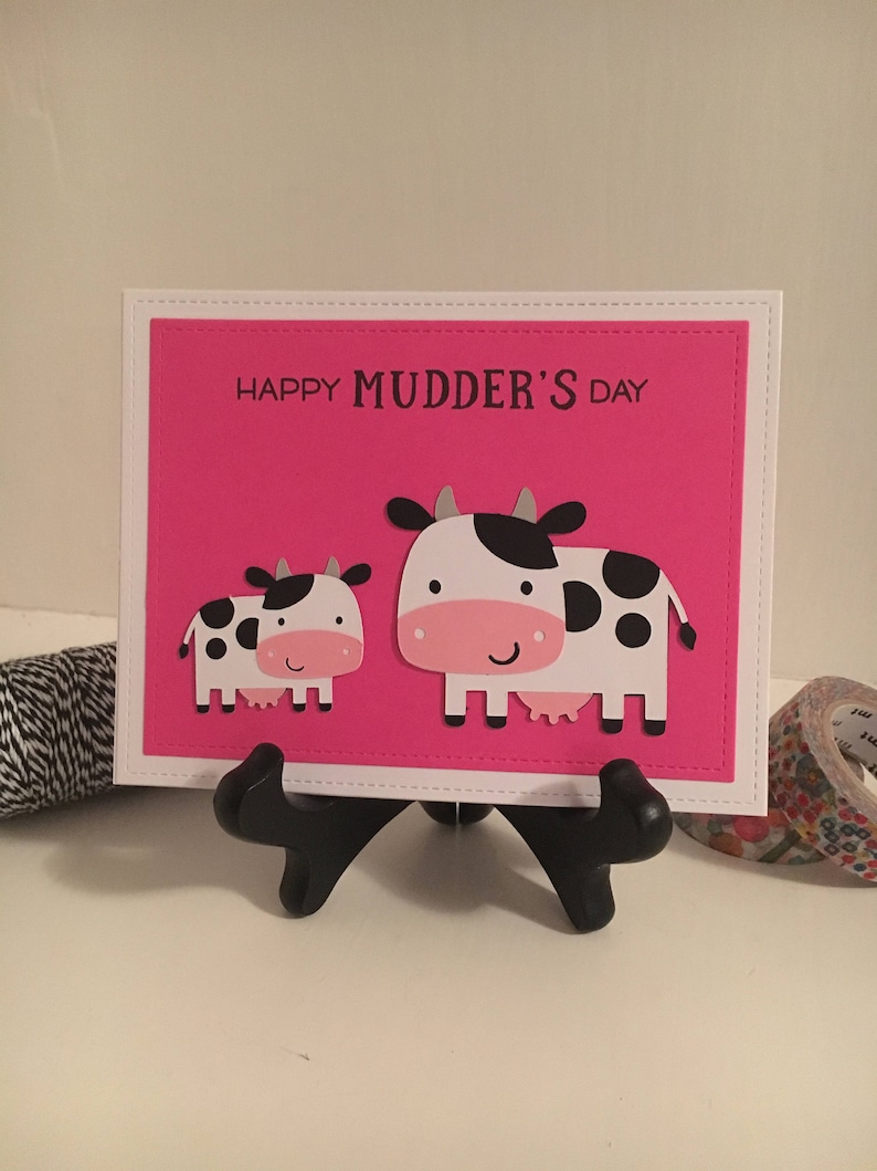 May include: A pink card with a white border and the text "Happy Mudder's Day" in black. Two cartoon cows are on the card, one larger than the other. The larger cow is white with black spots and the smaller cow is white with black spots.
