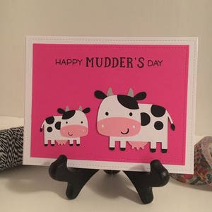 May include: A pink card with a white border and the text "Happy Mudder's Day". Two black and white cows are on the card, one is larger than the other.