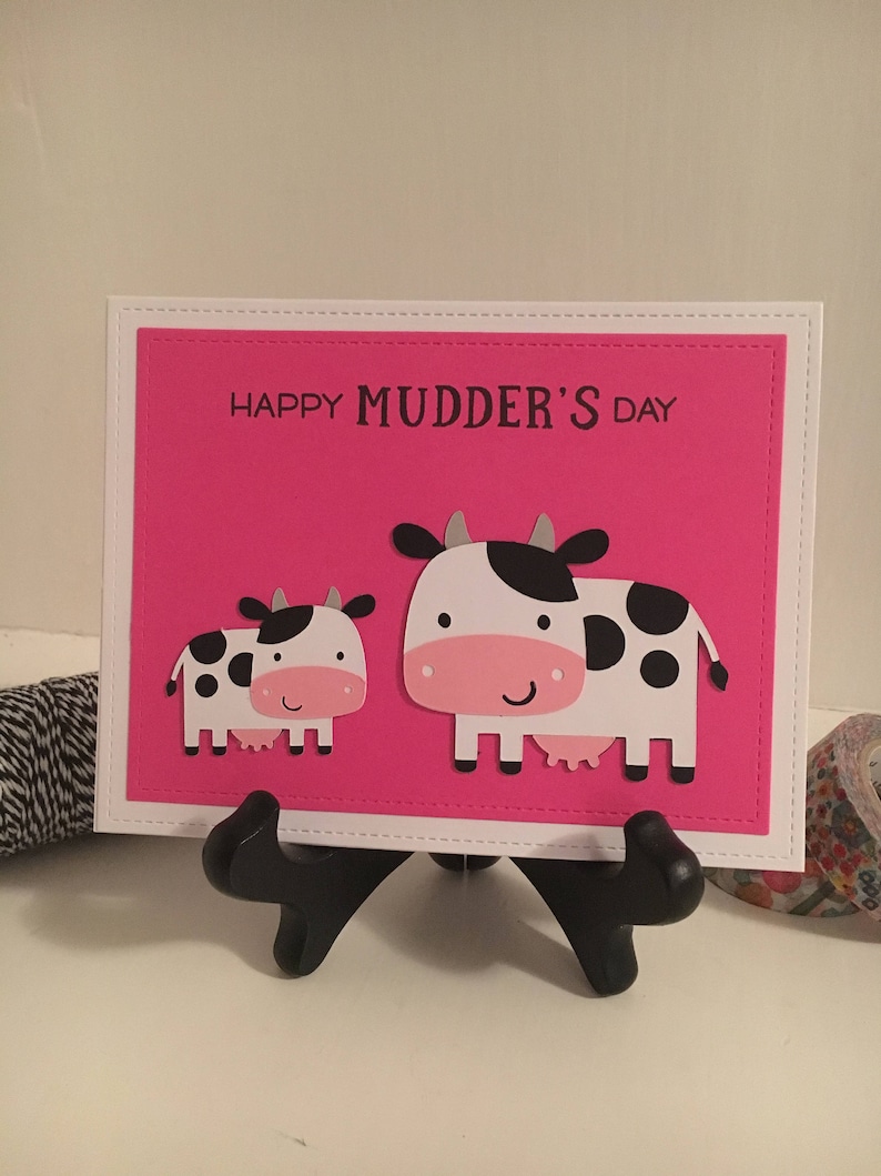 May include: A pink card with a white border and the text "Happy Mudder's Day". Two black and white cows are on the card, one is larger than the other.
