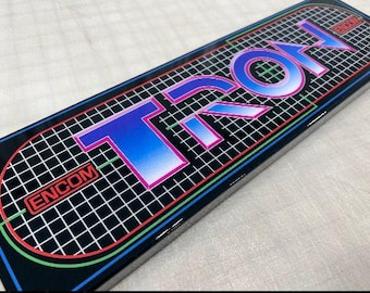 May include: A black and green game cartridge with the word "TRON" in a blue, pink, and white gradient on a black grid background. The text "ENCOM" is also visible in the lower left corner.