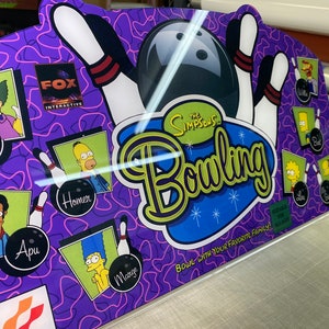 Könnte beinhalten: Ein lila-schwarzes Bowlingspiel mit Figuren aus den Simpsons. Das Spiel beinhaltet eine Bowlingkugel, Pins und Figuren wie Homer, Marge, Krusty und Apu. Das Spiel heißt "The Simpsons Bowling" und enthält den Text "Bowle mit deiner Lieblingsfamilie!"