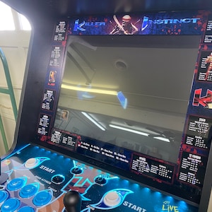 May include: A black arcade cabinet with a blue and white control panel. The cabinet has a large screen displaying the game "Killer Instinct". The game features characters like Jago, Cinder, and Fulgore. The cabinet has a start button and a high attack button.