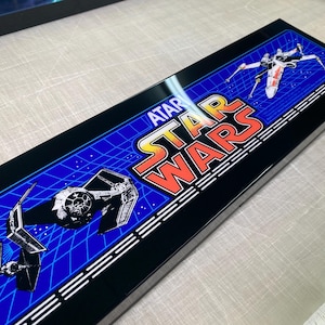 Puede incluir: Letrero rectangular negro con el logotipo de Atari Star Wars en naranja y amarillo. El fondo es azul con un patrón de cuadrícula e imágenes de naves espaciales X-wing y TIE fighter.