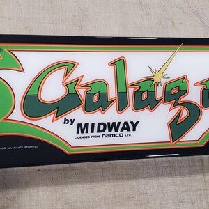 Könnte beinhalten: Ein schwarzer und weißer Arcade-Spiel-Marquee mit grünen und orangen Akzenten. Der Text auf dem Marquee lautet "Galaga by Midway Licensed from Namco Ltd. a Bally Co."
