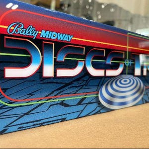 May include: A blue and red pinball machine cabinet with the text "Bally MIDWAY DISCS OF IRON" on the front. The cabinet has a blue and white striped disc on the front.