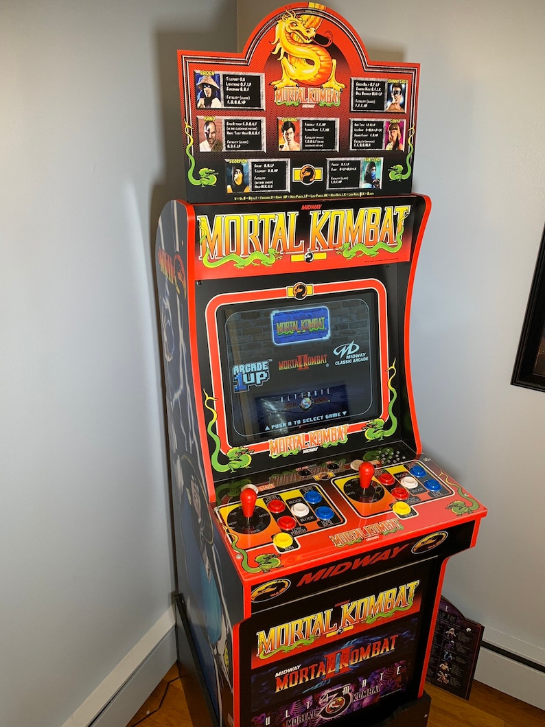 May include: A red and black arcade cabinet with the words "Mortal Kombat 2" on the front. The cabinet has a dragon design on the top and a screen that displays the game's title.