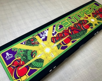 May include: A black and green arcade game control panel with the word "CENTIPEDE" in yellow letters. The panel features a green centipede, red and yellow mushrooms, and a yellow starburst.