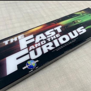 May include: A black and white rectangular sign with the text "The Fast and the Furious" in white. The Universal logo is in the bottom left corner. The sign has a green and yellow background with a blurry image of a green car.